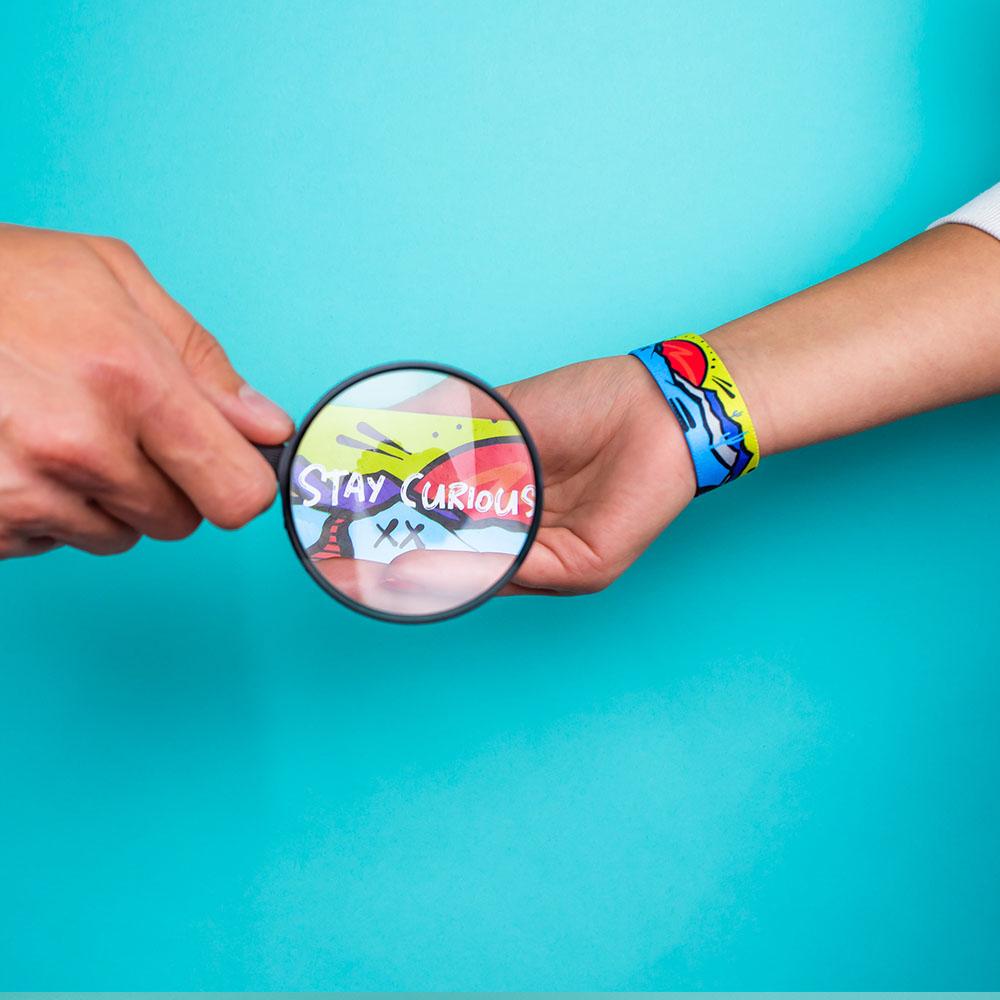 Studio image of hand holding a magnified glass over Stay Curious card and a Stay Curious on 1 wrist