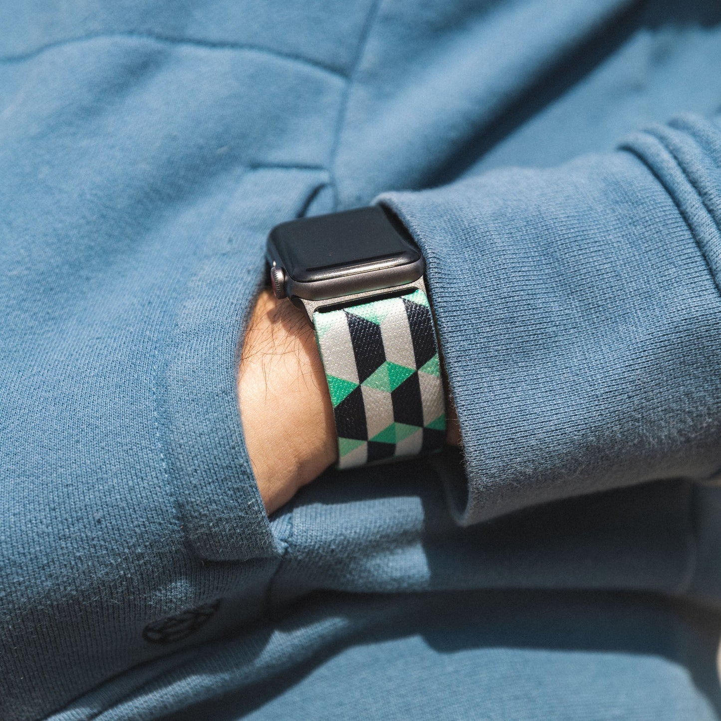 This is a lifestyle photo of a person wearing the watchband on an Apple watch. They are wearing a blue hoodie and have their hand inside the pocket. 