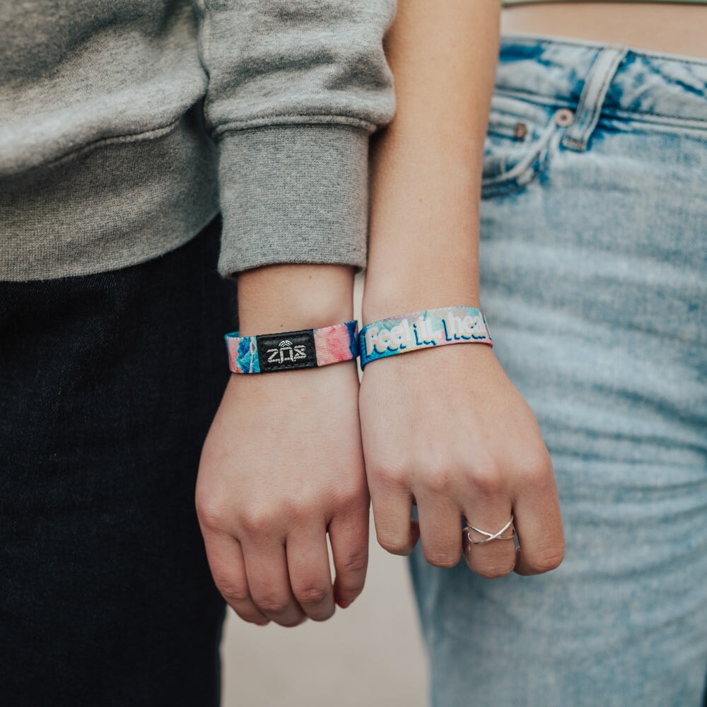 Two people stand side by side, each wearing a colorful bracelet from the Self-Care Pack. One wristband reads "Feel In." Both are casually dressed in jeans and sweatshirts, reflecting the playful self-care vibe of the set.