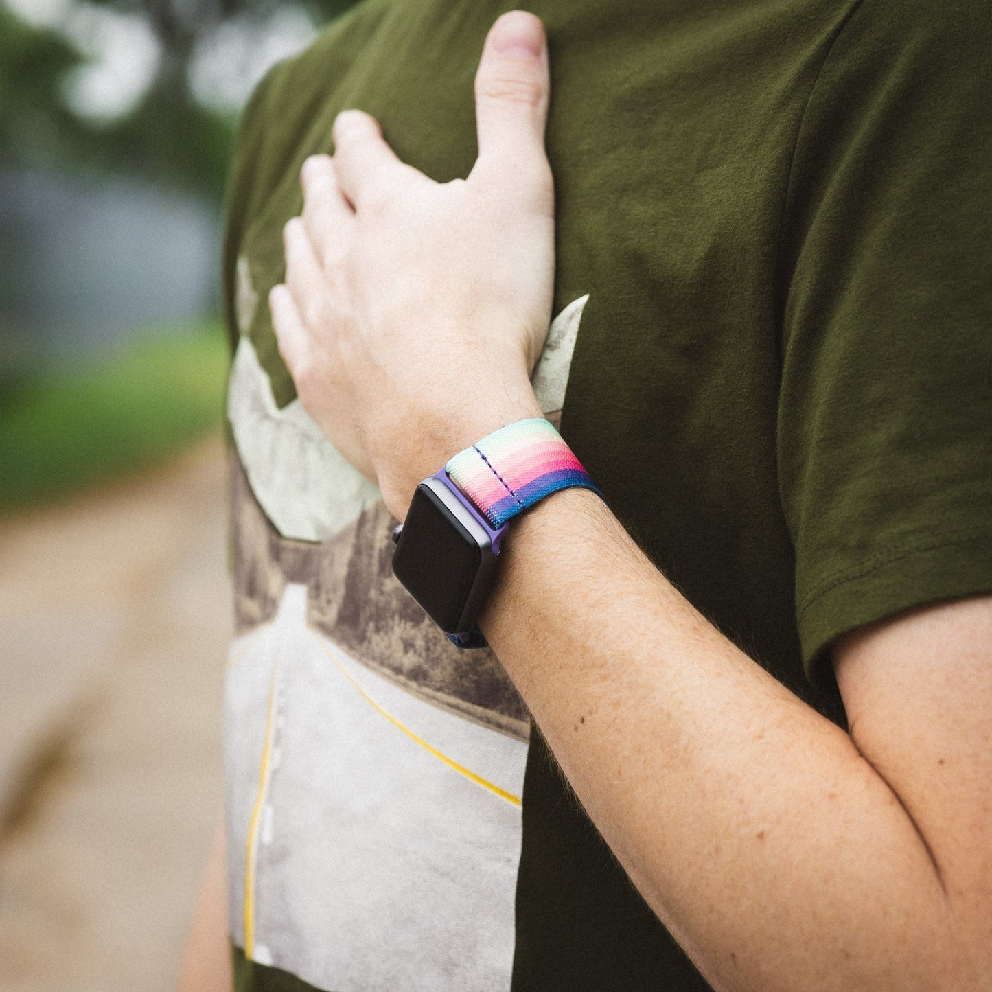 This is a lifestyle photo of a person wearing the watchband on an Apple watch. They have on a green tshirt and have their hand on their heart. 