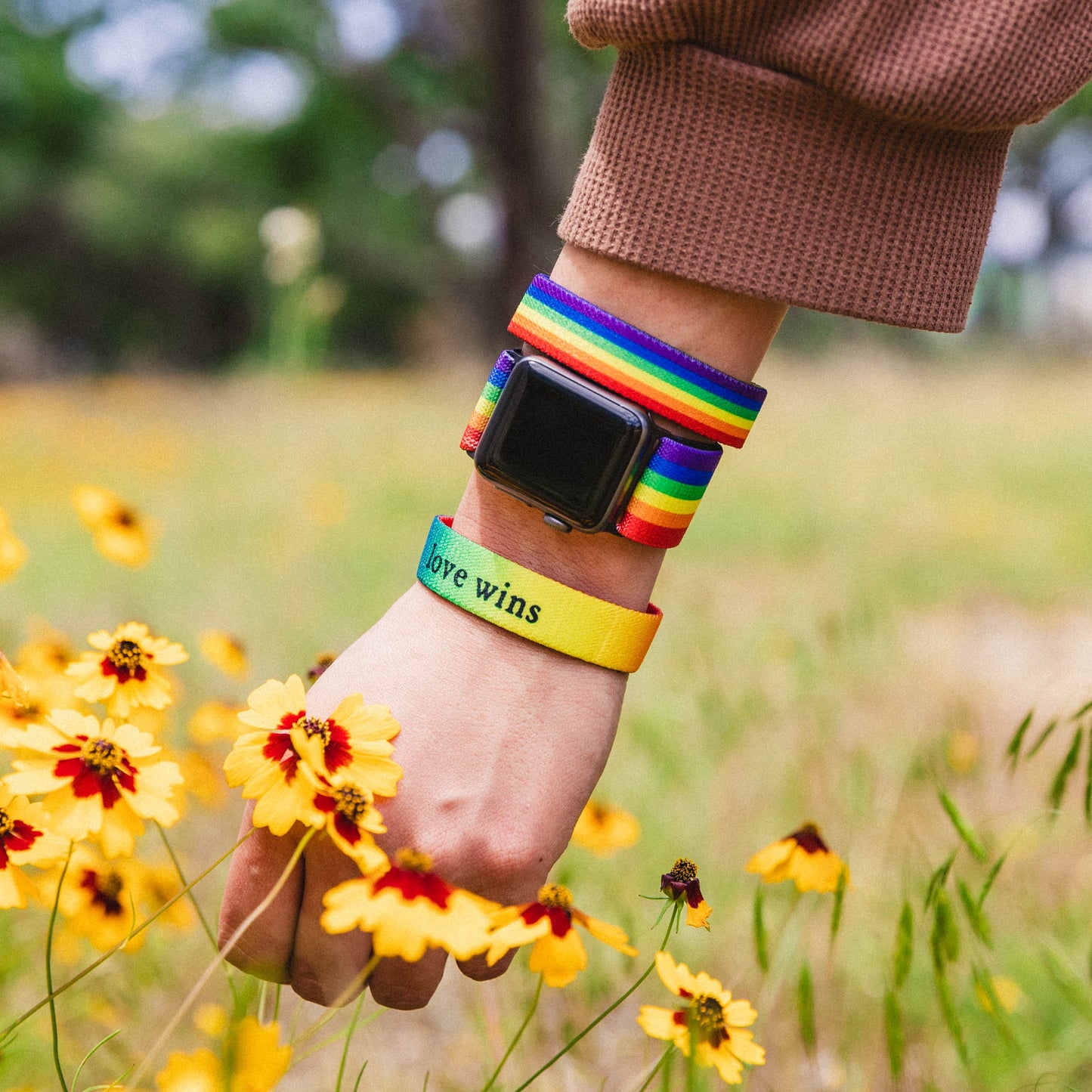 Lifestyle photo of someone wearing the Love Wins watchband. They have a matching Love Wins wristband on either side of the watch. One side is the design, one side is the quote. They are walking through a field of yellow and red wildflowers.