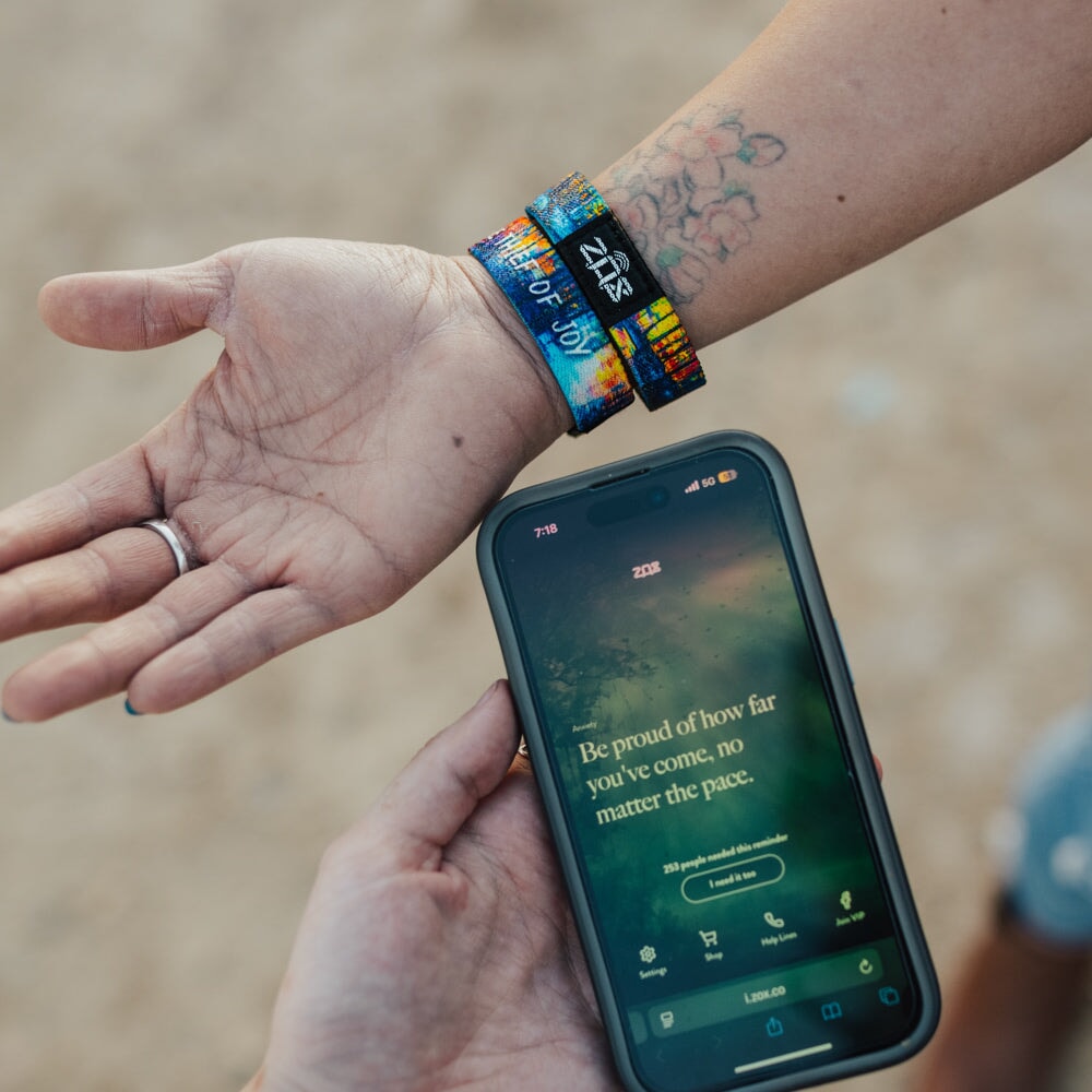 Wearing colorful festival wristbands and a floral tattoo, a person shows Self-Care Pack affirmation bracelets, while another displays an interactive affirmation on their phone: "Be proud of how far you've come, no matter the pace.
