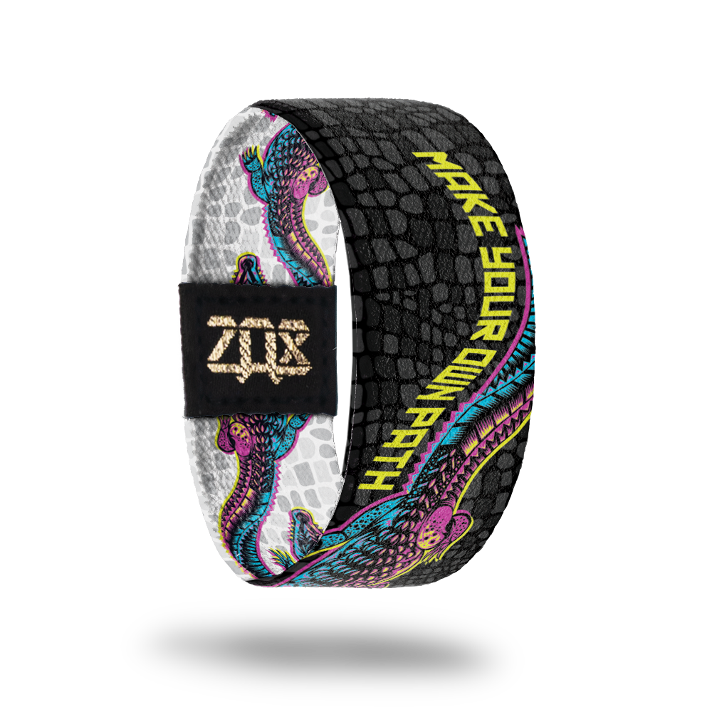Inside product photo of Make your Own path. An alligator that is pink, yellow and blue  is shown moving its tail and the words Make Your Own Path flow with the tail. The background is black alligator scales.
