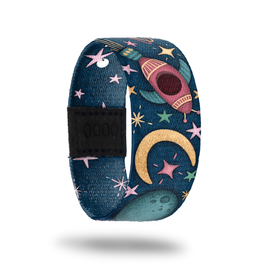 Wristband strap with a dark blue base of space. It has pastel colored stars and bursts with cartoon space ships flying through space. The inside is bluw with all light pink stars and reads One In A Lifetime. This comes with a matching lapel pin and a collector's box. 