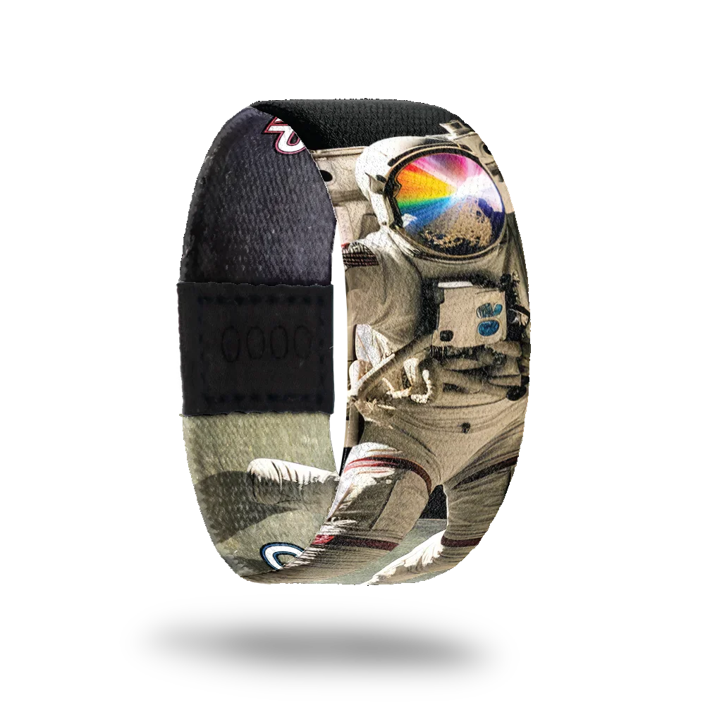 Wristband strap with a black base. A realistic black and white photo of an astronaut jumping on the moon. His face shield is reflecting off a rock and rays of rainbow colors. The inside is the same and reads One Giant Leap.