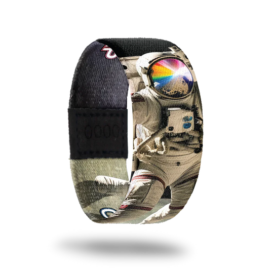 Wristband strap with a black base. A realistic black and white photo of an astronaut jumping on the moon. His face shield is reflecting off a rock and rays of rainbow colors. The inside is the same and reads One Giant Leap.