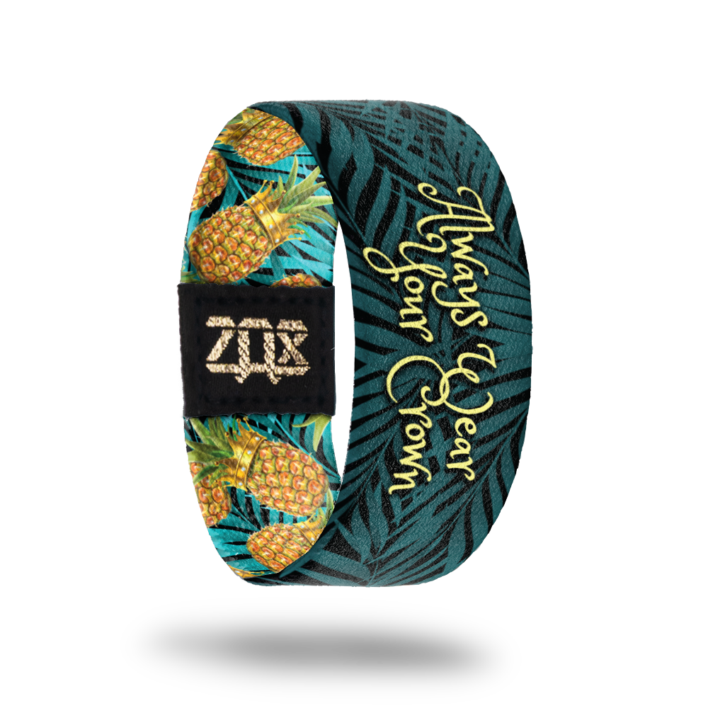 Inside of elastic wristband that says Always Wear your Crown in a light yellow font. Dark blue/green plants are in the background