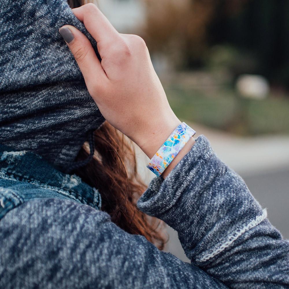 Lifestyle photo of hand grasping hood of jacket wearing dream big single showing outside design with a white geometric design overlaying a blue, purple, and yellow watercolored background 