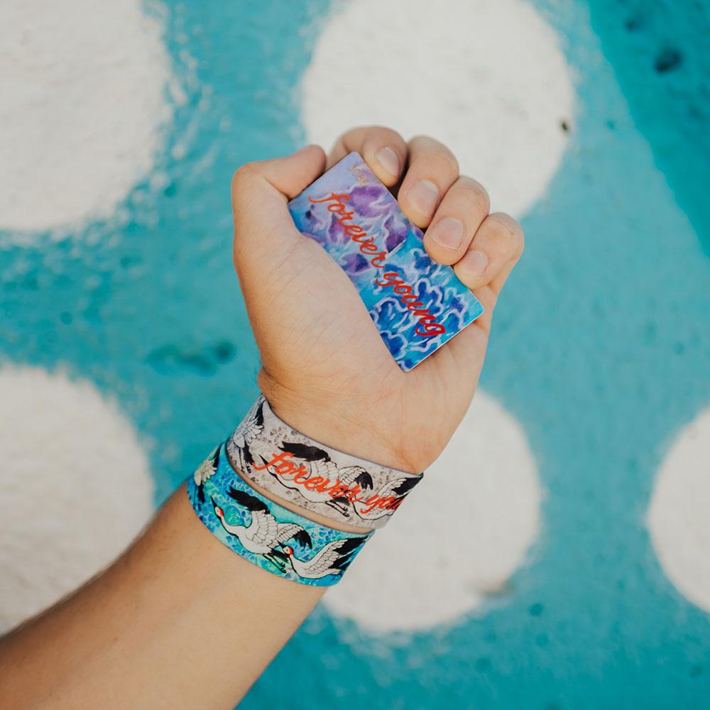 Lifestyle image of hand holding card that says Forever Young and on their wrist is 2 Forever Young straps