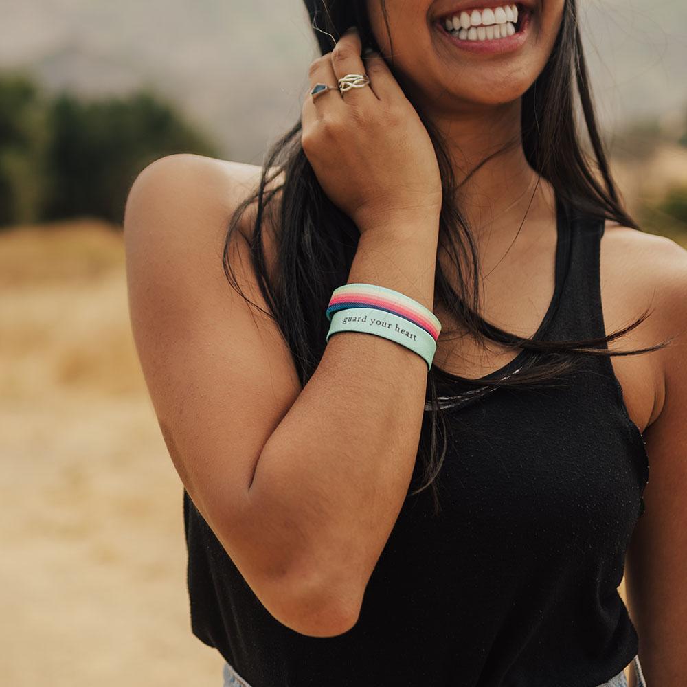 Lifestyle photo of girl wearing two guard your heart singles showing the outside design of guard your heart with mint, green, peach, pink, red, maroon, purple, blue rainbow design above another showing the  inside design with black text guard your heart with varying background in mint, green, peach, pink, red, maroon, purple, or blue