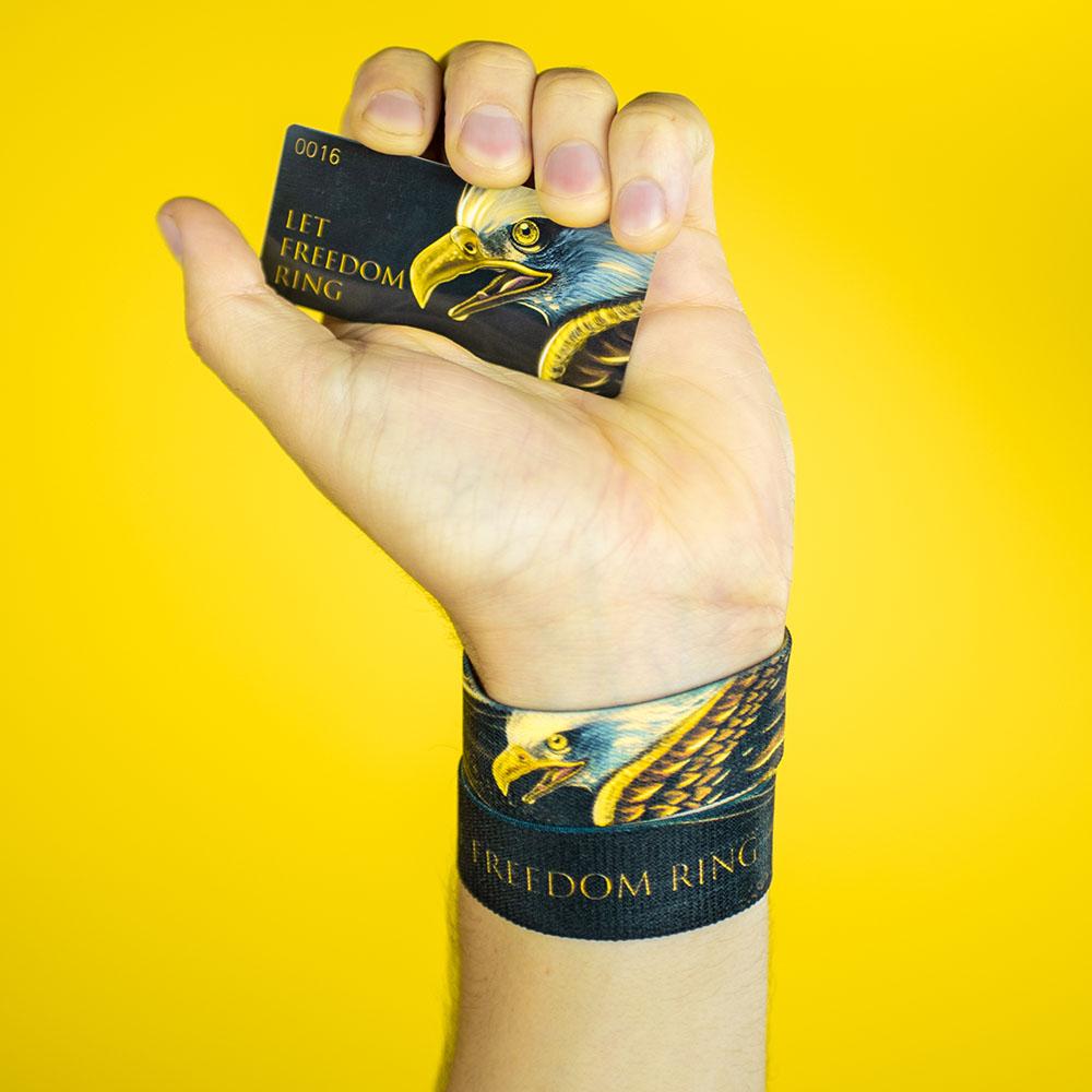 Studio Image of hand holding card that says Let Freedom Ring with a gold and white eagle head on the right side of the card and 2 Let Freedom Ring straps on their wrist