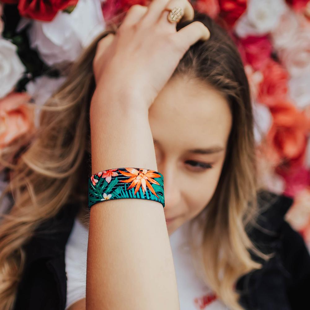 Lifestyle image of woman in front of a background of flowers and is covering her face with her arm which has Make It Happen on her wrist