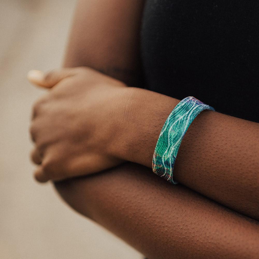 Lifestyle photo up close of a wrist showing the outside design of rise above it a white wave design over a green, pink, purple and orange watercolor background
