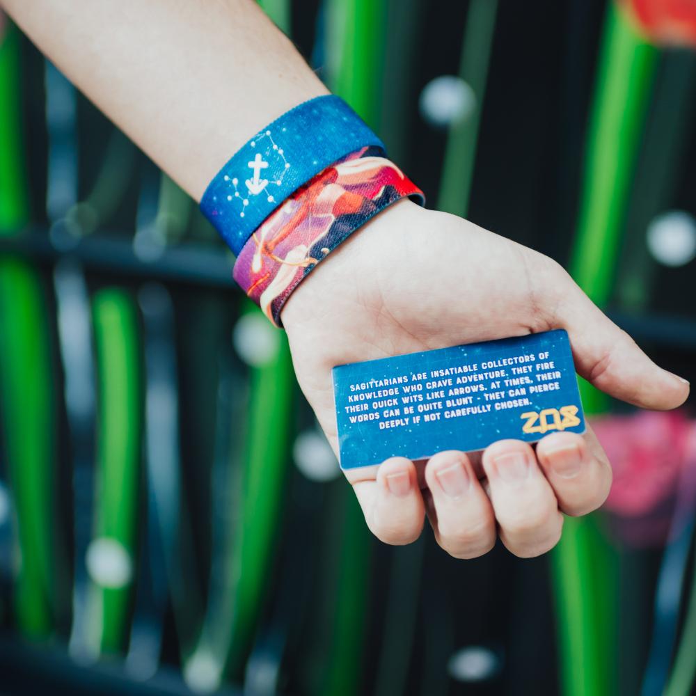 Lifestyle close up image of a hand holding collectable card that comes with Sagittarius strap and 2 Sagittarius straps are on their wrist